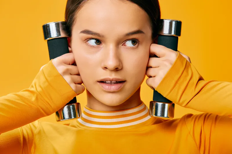 Fitness woman in a bright orange sweater uses dumbbells at shoulder height, focusing on form and strength. Vibrant orange studio backdrop emphasizes athletic energy and sport lifestyle in a clean,