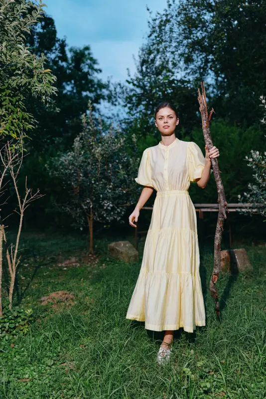 A woman in a pale yellow dress stands in a green garden holding a tall stick, calm and poised, soft sunlight filters through trees creating a peaceful outdoor scene