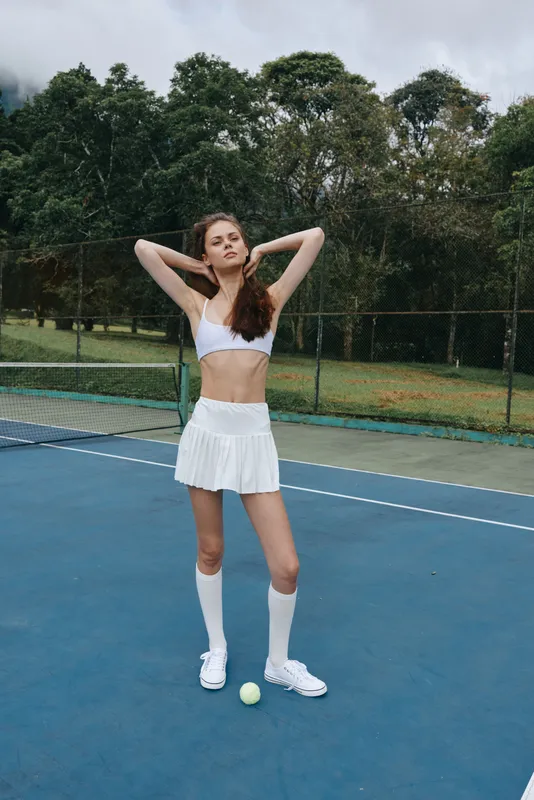 Athletic young woman in a white tennis outfit poses confidently on a tennis court, showcasing sportswear style and a vibrant outdoor atmosphere