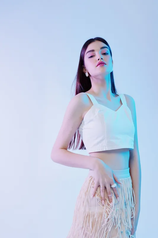 Young woman in stylish white crop top and fringed skirt poses confidently against a soft blue background. Concept of beauty, fashion, and modern style.