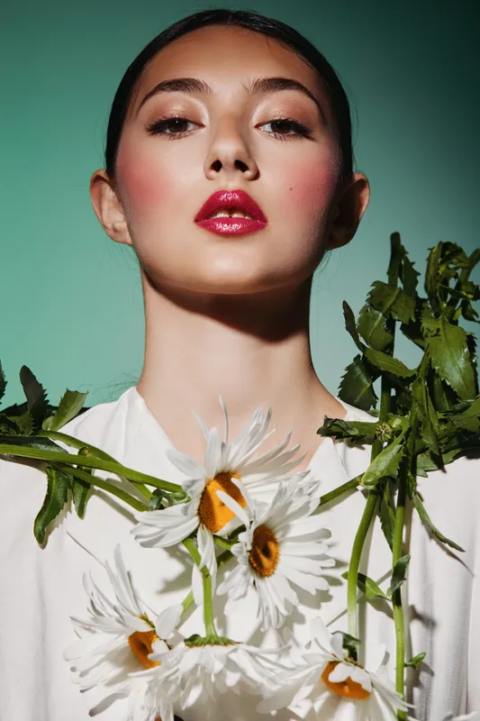 Portrait of a woman with glossy red lips and natural rosy blush. She holds fresh white daisies framed by green stems on a soft green background. Concept of creative beauty and fashion.