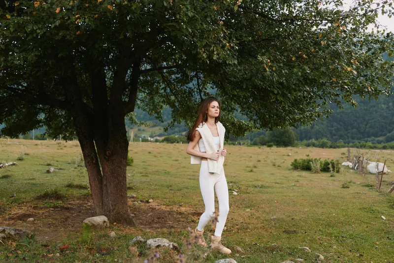 Young woman in white casual clothes walks thoughtfully near a large tree in a peaceful green meadow surrounded by hills under a cloudy sky.