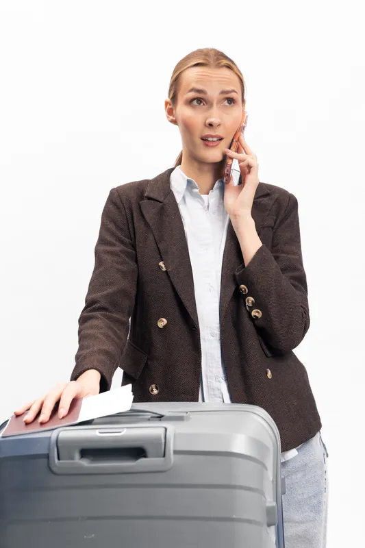 Young woman in brown blazer talks on phone while standing with large gray suitcase. White background emphasizes travel concept and business casual style. Expression shows concentration.