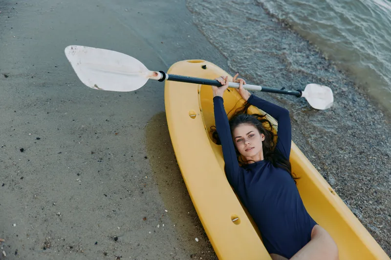 A person lounges in a yellow kayak on a quiet shoreline, holding a paddle overhead, sunlit water nearby, inviting a lazy yet adventurous coastal moment.