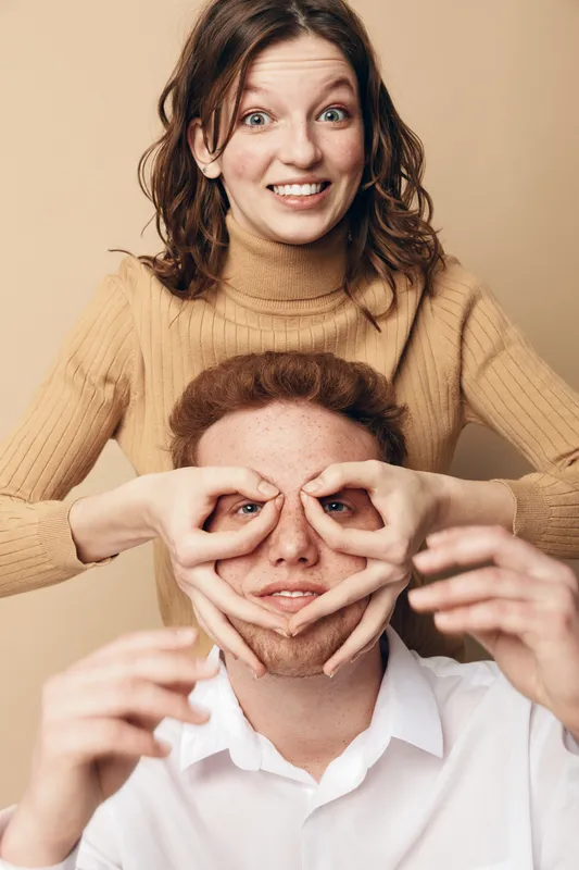 Smiling young woman in beige sweater playfully frames face of redhead man in white shirt with her hands. Studio portrait on beige background. Concept of friendship, fun, and creativity.