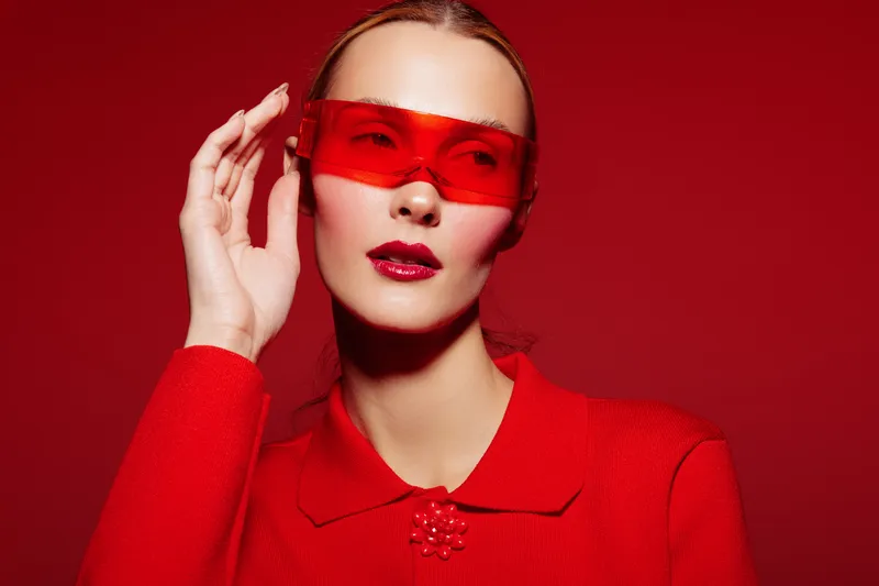 Fashionable woman in red outfit and red translucent sunglasses poses against red background. Concept of modern style, bold color, and confident female portrait in studio lighting.