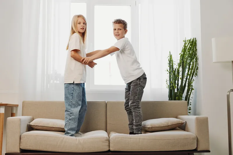 Two children stand on a beige sofa holding hands playfully in a bright living room with natural light. The smiling boy and girl enjoy a fun moment in casual clothes at home.