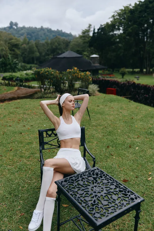 Young athletic woman in a white sports outfit enjoying a peaceful day in nature, surrounded by greenery, reflecting a healthy lifestyle and summer vibes
