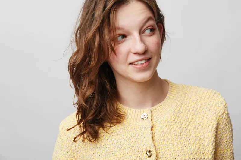 Young woman with long brown hair wearing yellow knitted sweater looks aside with slight smile on light grey background. Casual style, natural expression, portrait of relaxed female model.