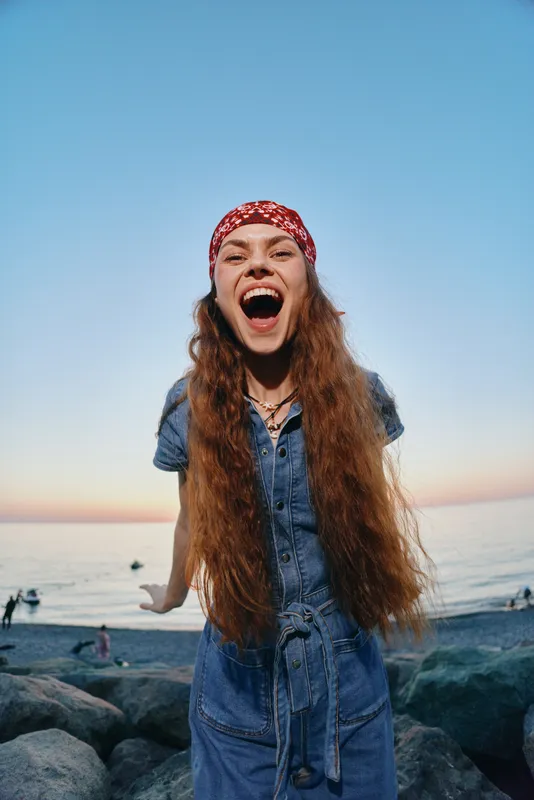 excited woman with long hair wearing red bandana and denim dress laughing loudly on rocky beach at sunset with clear sky