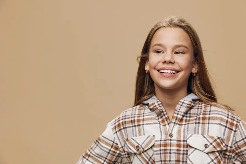 Girl smiling and looking to the side with light brown hair wearing plaid shirt in casual style against beige background. Youth, happiness, positive emotions and confident pose.