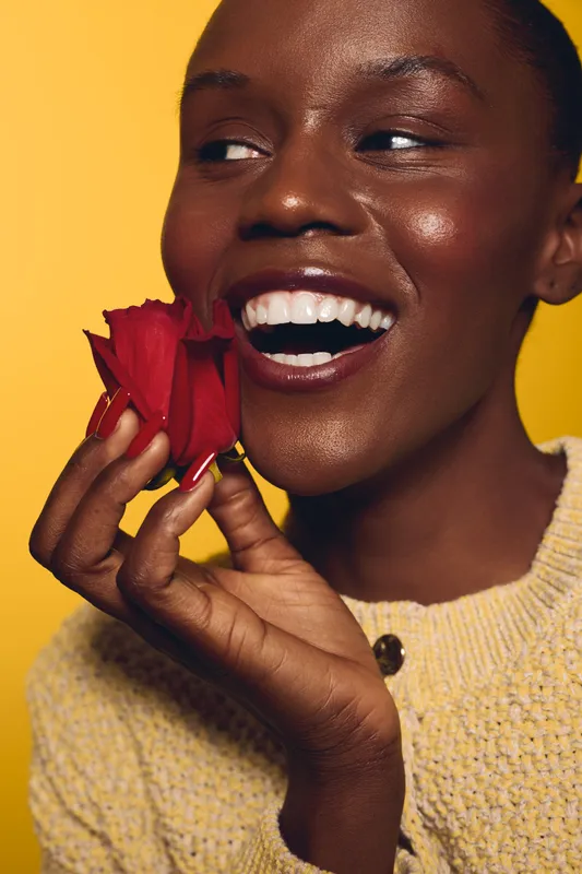 Close-up of a smiling African woman with dark skin holding red rose petals near her face. She wears a yellow sweater against a bright yellow background. Concept of beauty and joy.