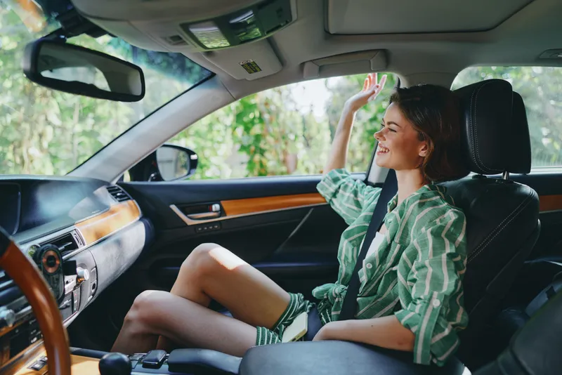 Young woman sits in a car interior, smiling and relaxed, enjoying a sunny road moment in casual attire.