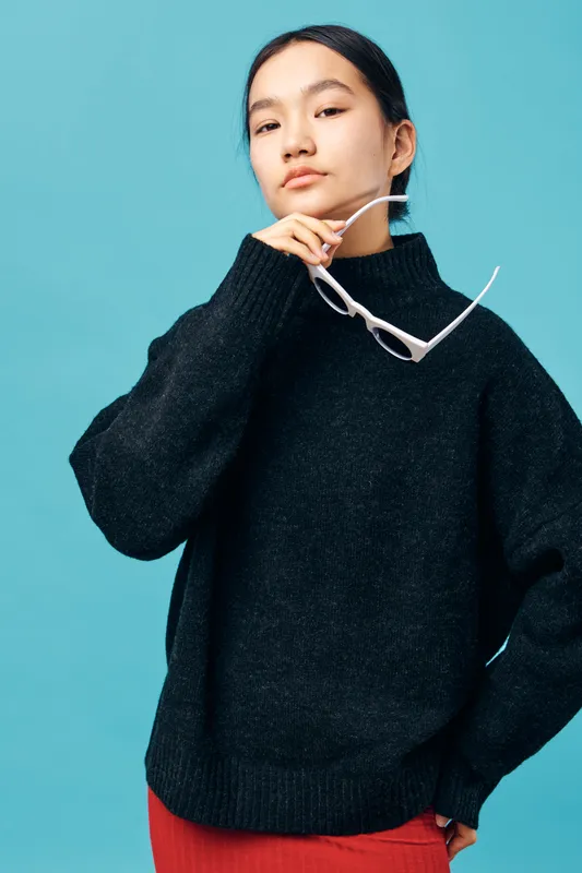 Young woman in black sweater holding white sunglasses posing confidently with hand near face against blue background. Fashion portrait of stylish casual outfit and natural makeup.