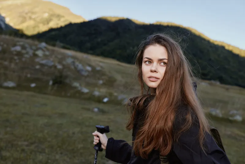 A thoughtful outdoor adventurer stands in a grassy field with distant hills. She holds a trekking pole and gazes toward the horizon, conveying calm, curiosity, and a sense of exploration.