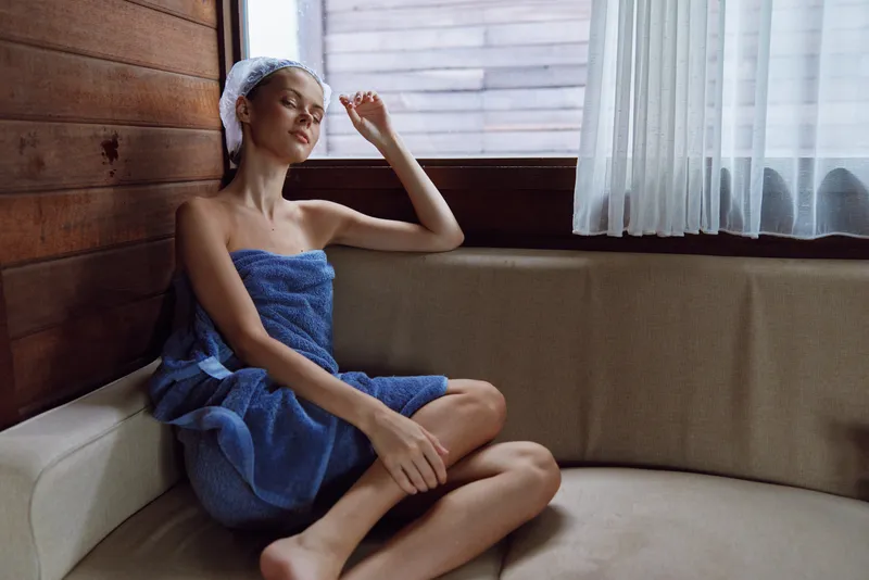 Relaxed woman wrapped in a blue towel sits on a beige couch in a rustic wooden room, enjoying a peaceful moment with natural light from a window.