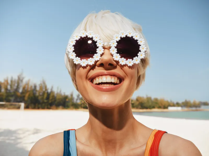 Bright sunlit portrait of a smiling person on a sunny beach wearing round flower sunglasses, capturing playful fashion and carefree summer vibe.