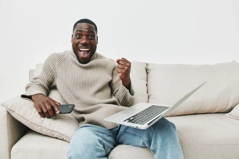 Young Black man celebrating success on a couch, wearing a cozy sweater and jeans, with a laptop and phone, expressing joy and excitement in a bright, modern living room