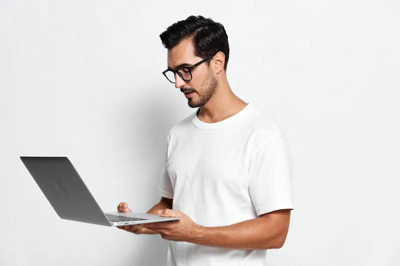 Focused handsome young man wearing glasses operates laptop while standing against plain light background. Technology and productivity concept.
