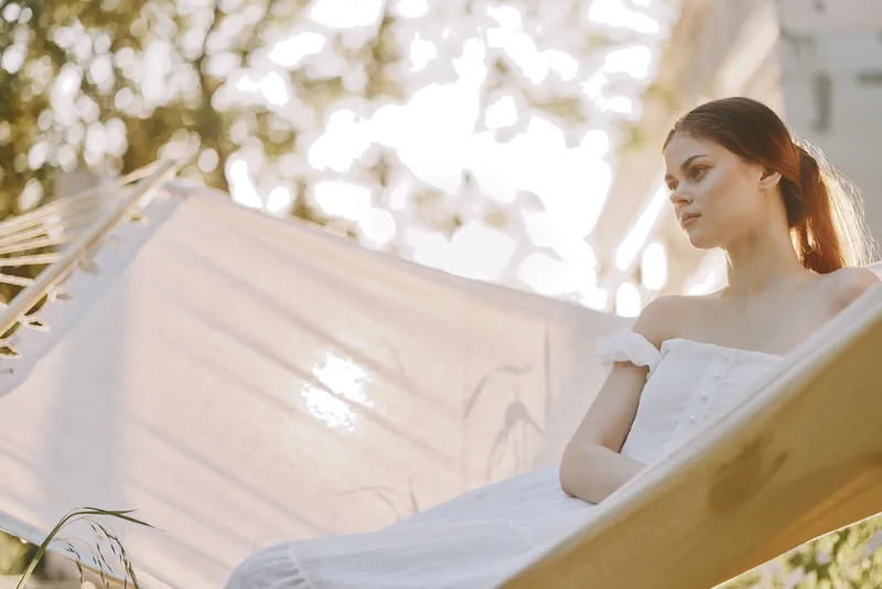Serene young woman in a white dress relaxing in a hammock, enjoying peaceful moments in nature during a sunny summer day