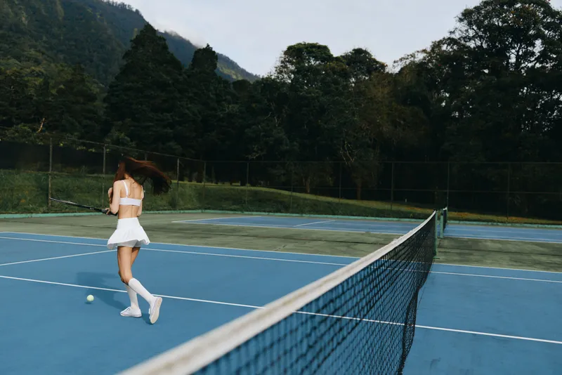 Young woman in white tennis outfit playing on blue outdoor court, showcasing athleticism and joy in a beautiful natural setting during daytime