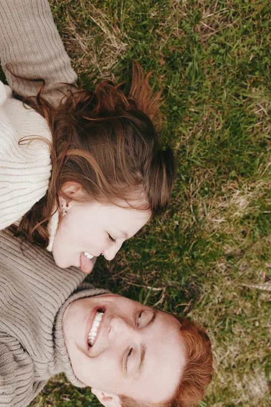 A young couple lies on the grass smiling and enjoying a playful moment together in cozy sweaters during a relaxed lifestyle outdoor scene.
