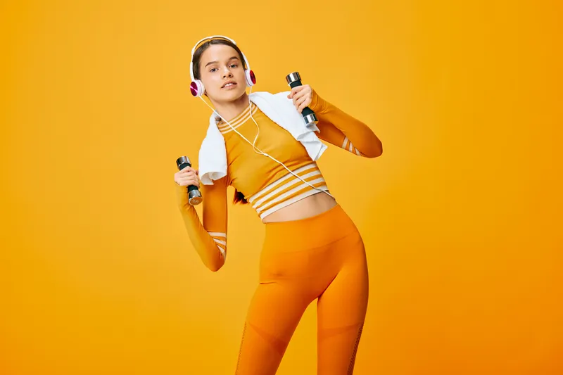 A woman fitness athlete in orange sportswear with headphones, lifting dumbbells against a bold orange backdrop, showcasing dynamic workout energy, focus and style in a studio scene.