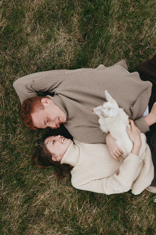 Cheerful couple outdoors, lying on grass, embracing a goat kid, sunlit moment, relaxed pose, soft sweaters, natural colors, intimate countryside lifestyle, warm breeze, gentle smiles, leafy backdrop,