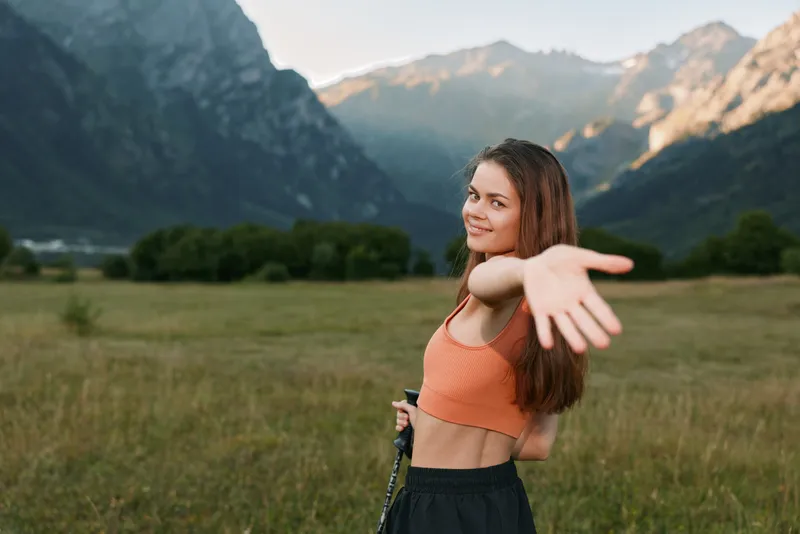 A cheerful young woman stands in a scenic valley, wearing a bright orange crop top. She smiles warmly and extends her arm toward the camera, creating a friendly, carefree outdoor mood.