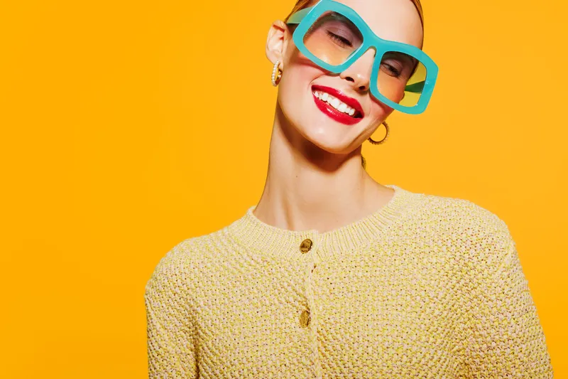 Smiling woman in oversized turquoise glasses and yellow sweater poses against bright orange background. Fashion portrait with vibrant colors and joyful expression, concept of style and confidence.