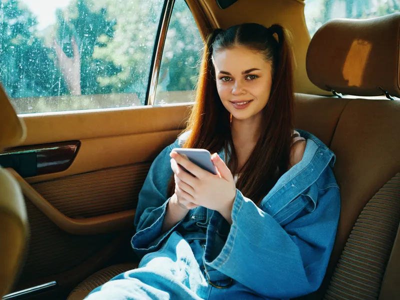 Young woman with long hair in casual denim clothes, smiling while using smartphone in a car, bright sunlight highlighting her features and relaxed atmosphere.