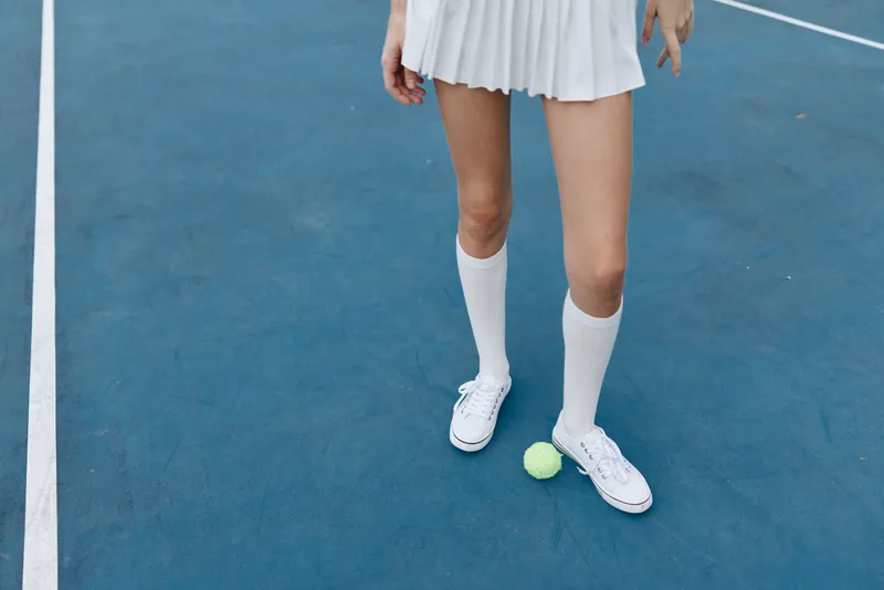 Young female tennis player in a white skirt and knee high socks standing on a blue court with a yellow tennis ball, showcasing athleticism and sportiness