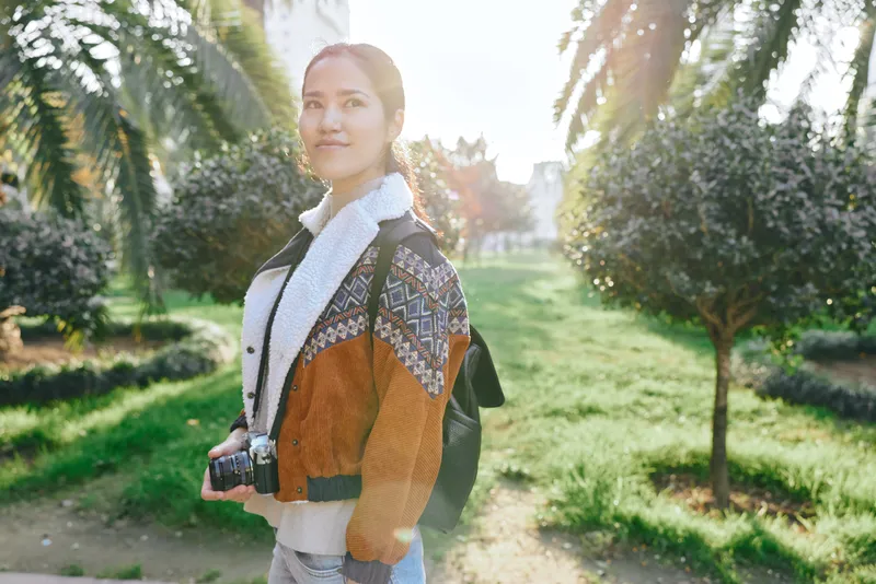 Woman with camera walks through a sunny park wearing a colorful knit jacket and backpack, capturing casual outdoor vibes and nature light during a relaxed photo session