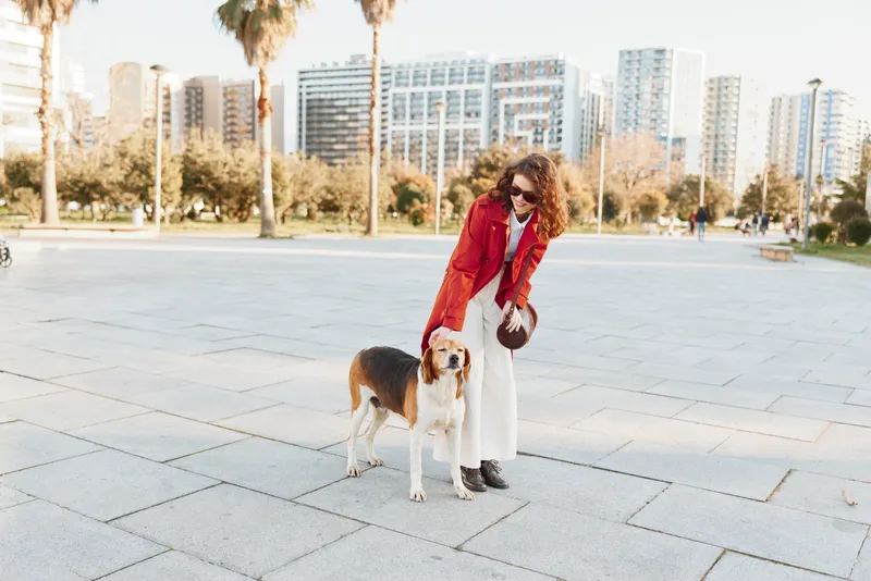 Smiling woman in a red jacket walks a beagle in a sunny city plaza, enjoying a casual urban stroll with her dog, her curly hair catching the light as modern buildings rise behind.