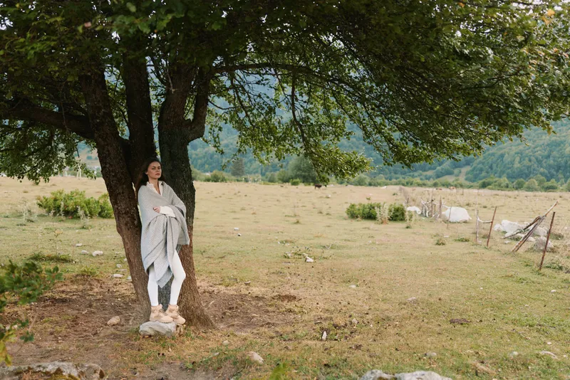A woman in a light wrap stands beside a large tree in an open countryside field, conveying calm, solitude, and a deep connection with nature during a peaceful day outdoors.