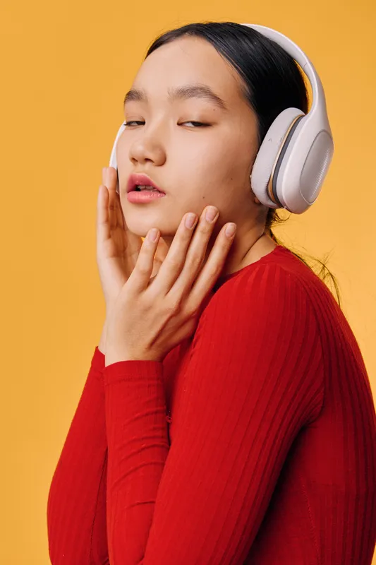 Young woman with headphones listening to music, wearing red sweater against yellow background. Relaxed person enjoying sound with closed eyes for entertainment or leisure.