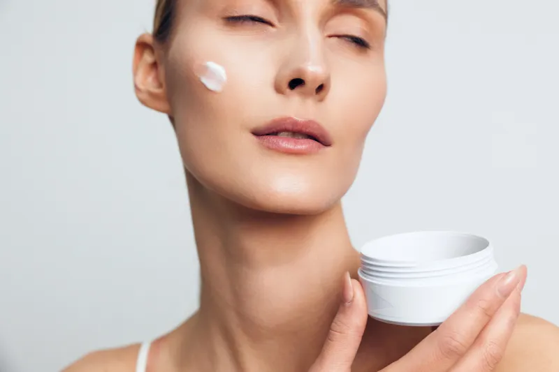 A young woman with brunette hair applies white face cream from a small white jar in a close up shot, against a plain light background, conveying a calm skincare moment.