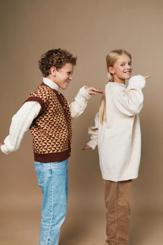 Two young children stand back to back, smiling and playful, posing in retro knitwear against a neutral backdrop, capturing carefree childhood fashion and friendship.