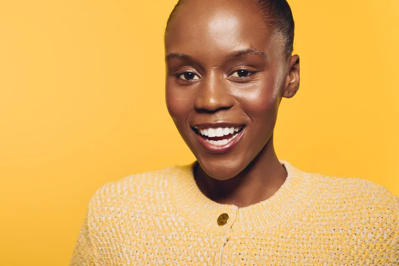 Smiling black African woman with dark skin wearing yellow sweater stands against yellow background. Portrait of happy female model expressing joy and confidence with natural beauty and bright mood.