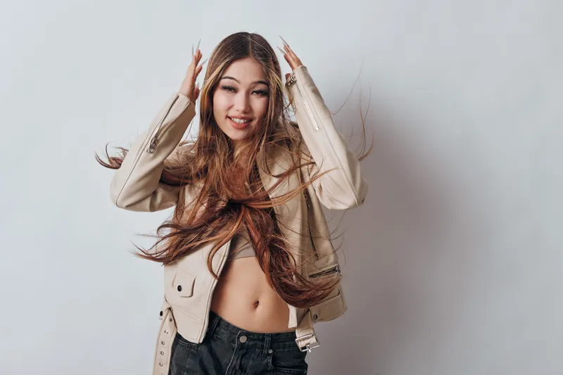 Young woman with long hair wearing beige jacket and black jeans standing against grey background. Portrait of fashionable female model posing with hands near head and smiling.