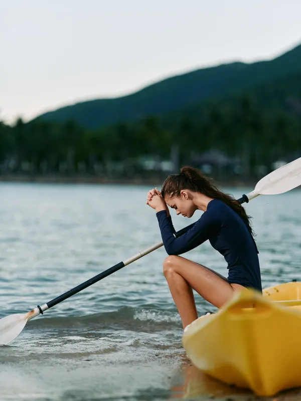 A lone paddler sits in a yellow kayak on a calm lake, wearing a dark wetsuit, head bowed, and holding an oar. Soft light and distant hills create a peaceful scene.