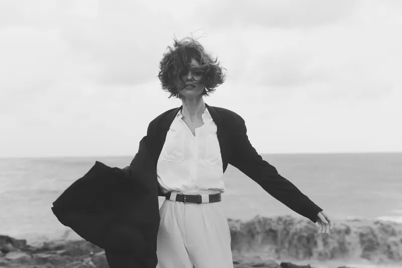 Fashion forward, windy seaside scene featuring a stylish figure in a blazer and white shirt, flowing coat, confident stance, ocean horizon, cloudy skies, movement and mood, contemporary elegance for