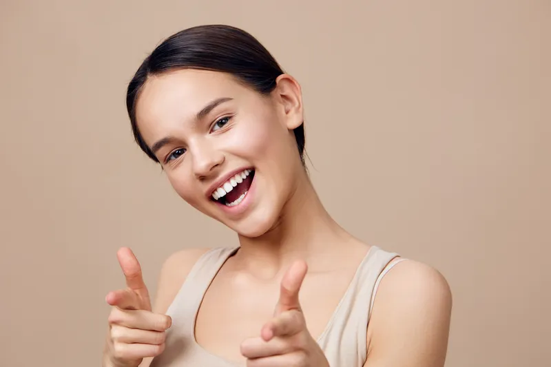 Cheerful young woman in casual beige outfit giving thumbs up and smiling, expressing positivity and confidence against a warm neutral background