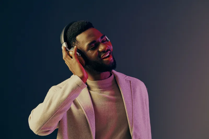 Young African man enjoying music with headphones, smiling and wearing a stylish sweater against a minimalist gradient background