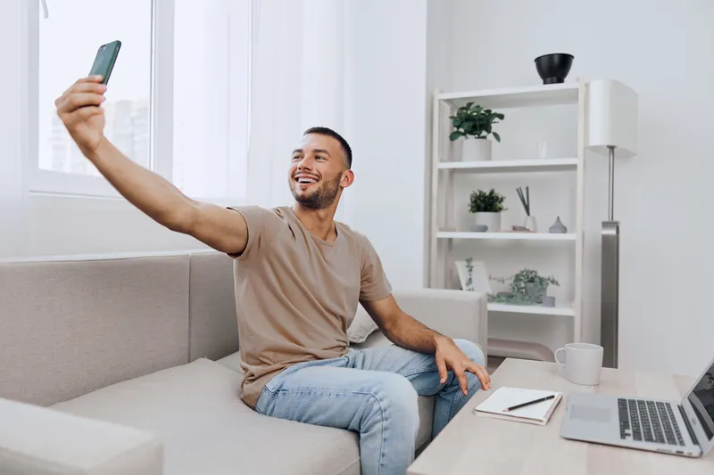 Smiling man taking a selfie at home, showcasing a cozy living room atmosphere with modern decor, capturing a moment of happiness and connection.