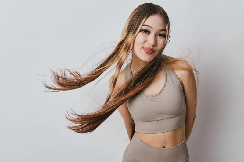Young woman with long hair wearing beige sportswear, posing smiling with hair flowing on light gray background in studio for fitness or casual portrait.
