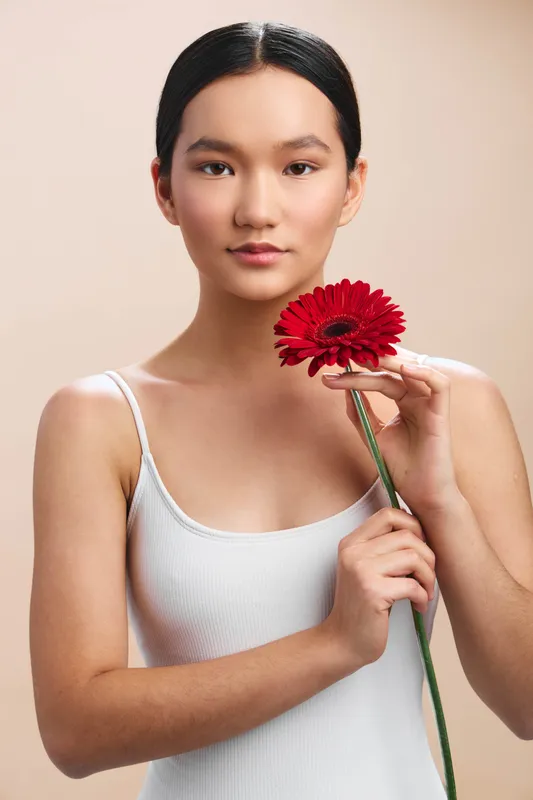 Asian woman in a simple studio setting gently holds a red flower, looking at the camera with calm confidence and poise, conveying elegance and serenity.