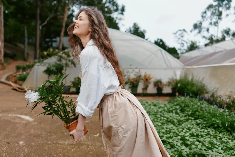 A joyful woman of European descent in casual attire holds a potted plant outdoors, showcasing her love for gardening against a natural backdrop