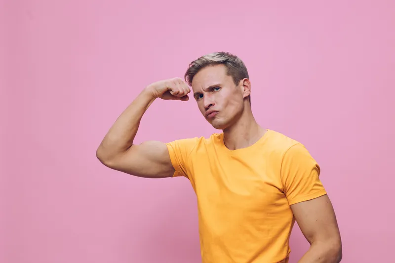 young caucasian man flex biceps strong fit muscle wearing orange t-shirt isolated pink background confident pose fitness concept