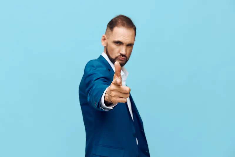 A confident business professional stands against a solid blue background, wearing a sharp suit and pointing toward the viewer, delivering a bold gesture to convey engagement and authority.